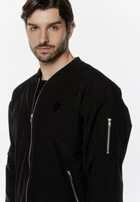 Czarna kurtka bomberka z gładkiego materiału, wyposażona w zamek błyskawiczny, ściągaczowy kołnierz oraz kieszeń na ramieniu z zamkiem błyskawicznym, z minimalnym logo.