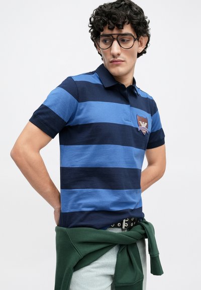 Πόλο πουκάμισο με εναλλασσόμενες μπλε ρίγες σε διάφορες αποχρώσεις, με σκούρο μπλε γιακά και ένα έμβλημα-μπαλωμα στην αριστερή πλευρά του στήθους.