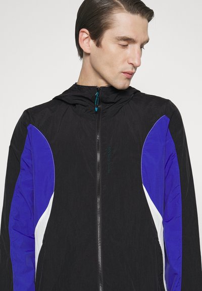 Schwarze Windjacke mit blauen und weißen Akzenten, Reißverschluss vorne und Kapuze. Leichter Stoff mit glatter Textur.