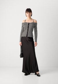 Haut noir et blanc à rayures, sans manches, avec détails en boutons, associé à une longue jupe noire fluide. Le modèle tient un sac noir et porte des chaussures noires.