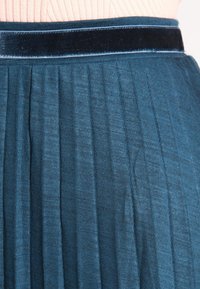 Gonna plissettata teal con cintura in velluto nero, tessuto texturizzato con righe verticali e un design morbido e fluido.