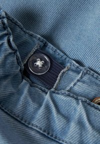 Tela de denim en un tono azul claro, con un botón de plástico transparente y un parche de tela azul oscuro para el cierre. Costura texturizada visible.