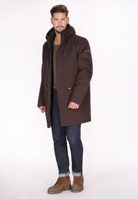 Brun parka jakke med hætte, frontlynlås, to sidelommer og snore. Bæres over en grøn skjorte og kombineret med mørke jeans og brune støvler.