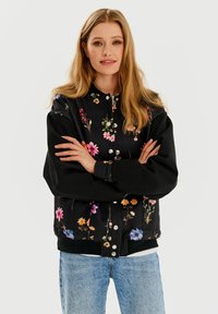 Czarna bomberka w kwiatowy wzór z kolorowymi motywami, zatrzaskami i luźnym krojem. Połączona z jasnoniebieskimi dżinsami.