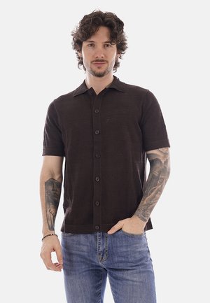 Giovane uomo con capelli ricci che indossa una camicia marrone scuro a maniche corte con bottoni e jeans blu, con tatuaggi su entrambe le braccia.