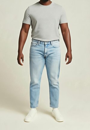 Homme portant un t-shirt gris clair à manches courtes, un jean bleu clair et des chaussures blanches sans lacets, debout devant un fond uni.