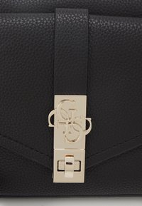 Černá kabelka z strukturované kůže s zlatým obdélníkovým sponou s propleteným písmem. Design zahrnuje šikmé prošívání.