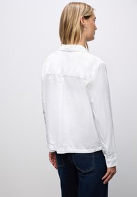 Camisa blanca de manga larga con cuello, detalle de yugo en la parte posterior y puños con botones. Textura suave, corte relajado y dobladillo ligeramente curvado.