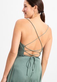 Satin-Kleid in gedämpftem Grün mit einem tiefen Rücken und einem Kreuzgurtdesign, ergänzt durch eine gebundene Akzentierung über einer glatten, flachen Textur.