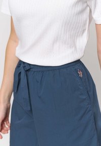 Tummansininen kevyet shortsit, joissa on joustava vyötärönauha ja kiristysnauha, sivussa pieni kirjailtu yksityiskohta. Yhdistetty valkoiseen teksturoituun toppiin.