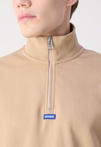 Pullover con zip di colore tan con colletto alto. Presenta una zip argentata e una patch del logo blu del marchio. Realizzato in un morbido tessuto testurizzato.
