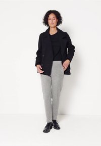 Zwarte wollen jas, getailleerde grijze checkbroeken, zwart top en zwarte schoenen. Model staat met haar handen langs haar lichaam tegen een witte achtergrond.