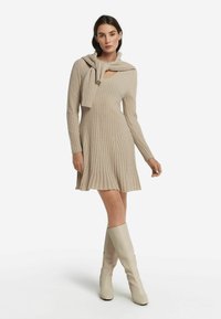 Beige geripptes Strickkleid mit langen Ärmeln, V-Ausschnitt und ausgestelltem Saum. Pullover über die Schultern drapiert. Model trägt knielange beige Stiefel.