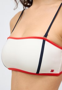 Λευκό πάνω μέρος μπικίνι με ναυτικού μπλε λουράκια και κόκκινο περίγραμμα. Ομαλή υφή, ορθογώνιο σχήμα, με ένα μικρό λογότυπο στην κάτω δεξιά γωνία.
