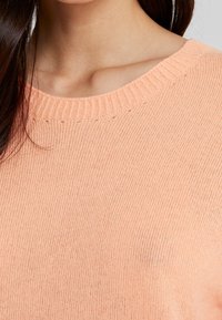 Ljus persikofärgad tröja, mjuk struktur, ribbad halsringning och korta ärmar. Närbild visar stickmönster och sömmar.