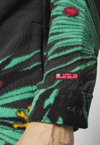 Gros plan sur une manche en polaire verte et noire à motif, avec un patch logo rouge et un poignet noir à boutons sur le bras d'une personne.