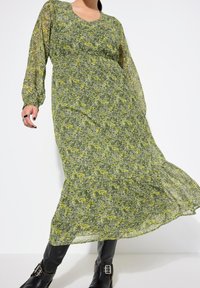 Abito lungo verde floreale con motivi gialli e bianchi, vita arricciata e maniche lunghe trasparenti. Stivali neri al ginocchio completano l'outfit.