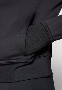 Détail d'un sweatshirt noir avec un tissu texturé, des poignets côtelés et une poche latérale intégrée dans le design. Finition en matériau lisse.