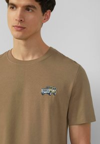 Mann trägt ein braunes Rundhalsausschnitt-T-Shirt mit einem kleinen gestickten gelben und schwarzen SUV-Design auf der linken Brust.