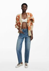 Beżowy sweter na drutach z pomarańczowym kwiatowym wzorem, biały krótki top i niebieskie dżinsy z wysokim stanem. Modelka nosi białe trampki.