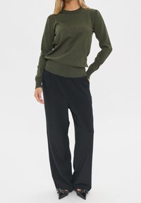 Olivgrön stickad tröja med långa ärmar, ribbade muddar och rund halsringning, kombinerad med löst sittande svarta byxor och spetsiga svarta klackskor.