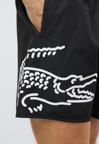 Μαύρα μαγιό σορτς με λευκό γραφικό κροκόδειλου στο αριστερό πόδι. Το υλικό είναι απαλό και γρήγορα στεγνώνει. Διαθέτουν πλαϊνές τσέπες.