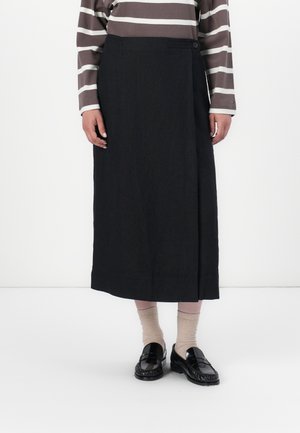 Personne portant une jupe midi noire, des chaussettes beiges, des mocassins noirs brillants et une chemise à manches longues rayée marron et blanche, debout devant un fond blanc.