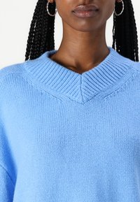 Lyseblå strikkegenser med ribbet V-halsdesign og strukturgarn. Ermene er vide og har drop-shoulder design.