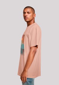 Helles rosa T-Shirt mit kurzen Ärmeln, das ein grafisches Design von Bergen und Bäumen in Teal und Orange zeigt. Weicher Stoff, lässige Passform.