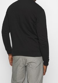 Sudadera negra de mangas largas, con puños y dobladillo acanalados, hecha de un material suave, vista desde la parte de atrás, combinada con pantalones claros de cuadros.