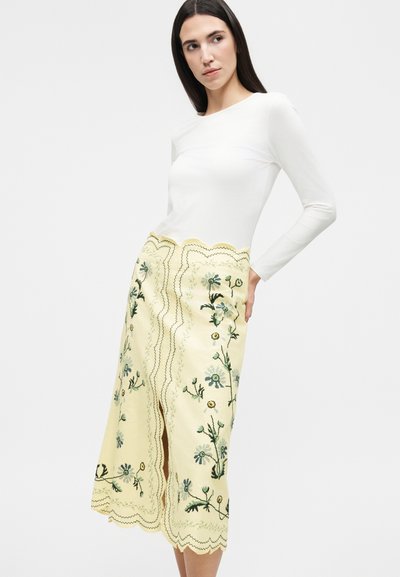 Γυναίκα που φοράει λευκό τοπ με μακριά μανίκια και κίτρινη μίντι φούστα με ψηλή μέση, κεντητά λουλουδάτα σχέδια και κυματιστές άκρες, ποζάροντας με τα χέρια πίσω από την πλάτη.