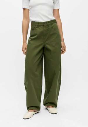Vrouw in olijfgroene, wijde, hoge taille broek, wit kort mouw shirtje en witte platte schoenen staand tegen een effen achtergrond.