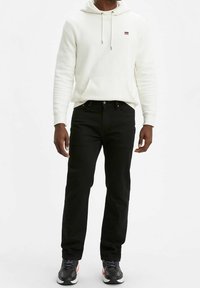 Sudadera blanca con bolsillo frontal, jeans negros y zapatillas deportivas negras. La sudadera tiene cordones y un pequeño logo en el pecho.