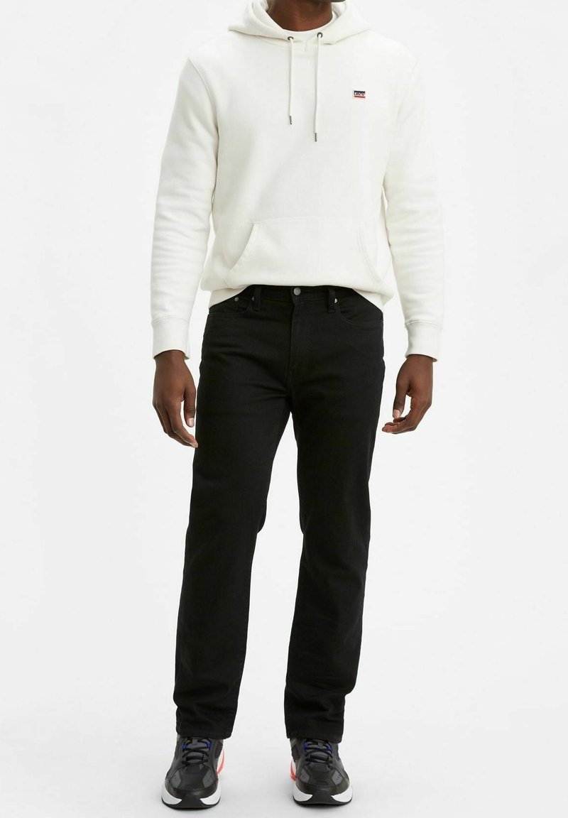 Sudadera blanca con bolsillo frontal, jeans negros y zapatillas deportivas negras. La sudadera tiene cordones y un pequeño logo en el pecho.