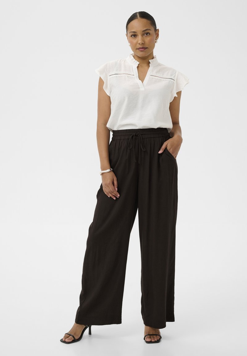 Femme portant un chemisier blanc à manches courtes à volants, pantalon large noir taille haute, talons ouverts noirs, debout avec une main dans la poche.