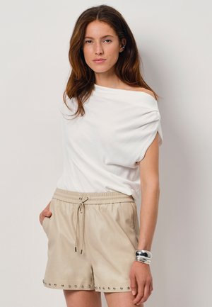 Witte top met draping en korte mouwen; beige shorts met elastische tailleband en zilveren studs aan de zoom; gladde stof; gedragen met een zilveren armband.
