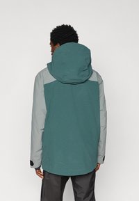 Sötét göndör hajú férfi, aki teal és világosszürke kapucnis dzsekit visel, háttal áll egy sima fehér háttér előtt.