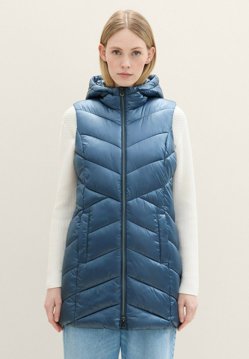 Modrý puff vest s chevron vzorom, dizajnom s kapucňou a predným zipsom. Nosený cez biely textúrovaný sveter. Pod tým sú svetlé denimové džínsy.
