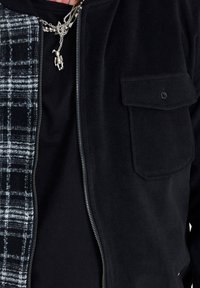 Personne portant une veste noire à fermeture éclair avec une doublure intérieure à carreaux et un collier argenté avec un petit pendentif en forme d'ours en peluche suspendu.