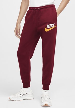 Homme portant un pantalon de survêtement Nike bordeaux avec un logo jaune et des baskets Nike blanches avec un swoosh noir, debout sur un fond uni.