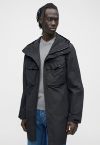 Černá bunda s kapucí, předním zipem a dvěma kapsami na prsou. Texturovaná tkanina, volný střih, vrstvená přes šedý svetr s výstřihem do V a modré džíny.