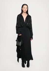 Black long-sleeve blouse features a deep V-neck and shiny fabric. Paired with a flowing black skirt and a studded handbag. Black heeled boots included.