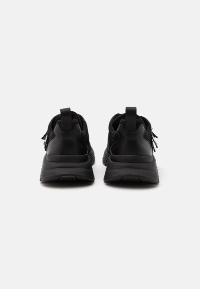 Paire de chaussures de sport noires vues de dos sur un fond clair uni, avec languettes à tirer et semelles texturées.