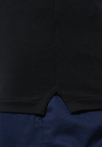 Schwarzes Polohemd aus strukturiertem Stoff mit einem Belüftungsabschluss. Trägt es über dunkelblauer Hose, die ein sauberes und einfaches Design zeigt.