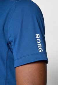 Mėlyna trumpomis rankovėmis marškinėliai su tekstūruotu audiniu. Kairiajame rankovėje yra baltas "BORG" logotipas. Siūlės matomos palei siūles.