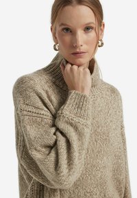 Beiger Strickpullover mit hohem Kragen, aus strukturiertem Stoff und mit gerippten Bündchen. Akzente setzen ein detailliertes Strickmuster und eine lockere Passform.