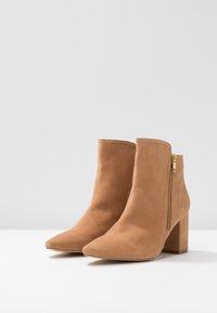 Botas de ante hasta el tobillo en color marrón claro con un alto tacón cuadrado, punta afilada y un detalle decorativo de cremallera dorada en el lado exterior.