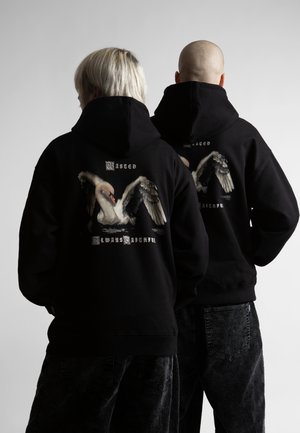 Dvaja ľudia, jeden s blond vlasmi a druhý plešatý, stoja vedľa seba oblečení v čiernych mikinách s kapucňou s veľkým obrázkom labute a textom na chrbte.