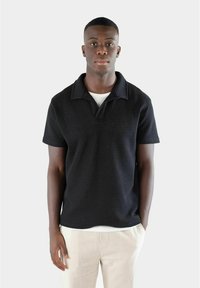 Svart kortärmad pikétröja med ett texturerat mönster och nedfällbar krage, bärs över en vit T-shirt och matchas med ljusa byxor.