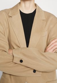 Persona con un blazer beige sobre una camiseta negra de canalé, con los brazos cruzados sobre el pecho, recortada del cuello a la cintura sobre un fondo blanco.